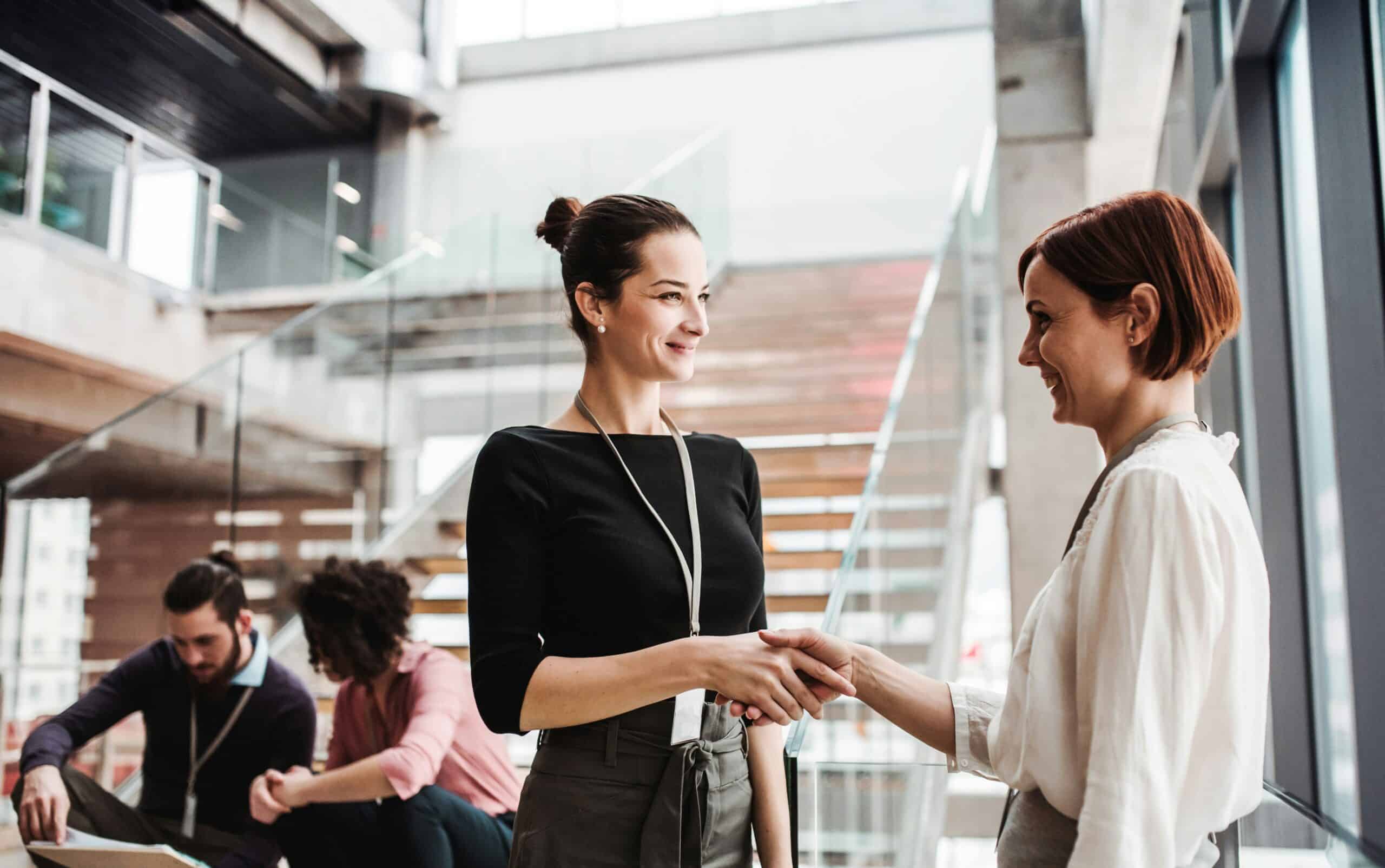 Two professional women smiling and shaking hands in a modern, sunlit office atrium with stairs in the background.