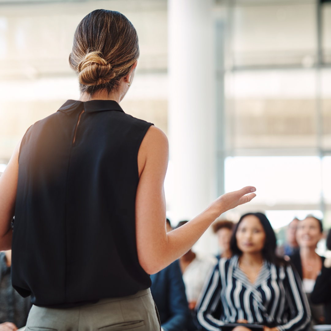 Speaker addressing an audience, viewed from behind, with attendees listening attentively in a bright, modern setting.